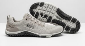 A pair of light grey Xero Shoes with branding on the side; one shoe is upright, while the other on its side shows a black and white textured sole—designed for walking and all-day comfort with a visible tread pattern.