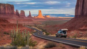 A winding road meanders through the Desert Southwest, passing striking red rock formations and mesas. An SUV with a roof box heads toward distant cliffs. Saguaro cacti punctuate the scene beneath soft clouds, highlighting Monument Valley's iconic road trip landscape.